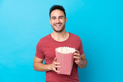 handsome man over isolated blue background holding a big bucket of popcorns